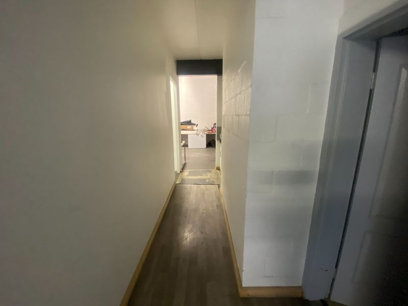 A narrow commercial hallway with laminate flooring and painted masonry walls, leading to an adjacent utility or storage area.