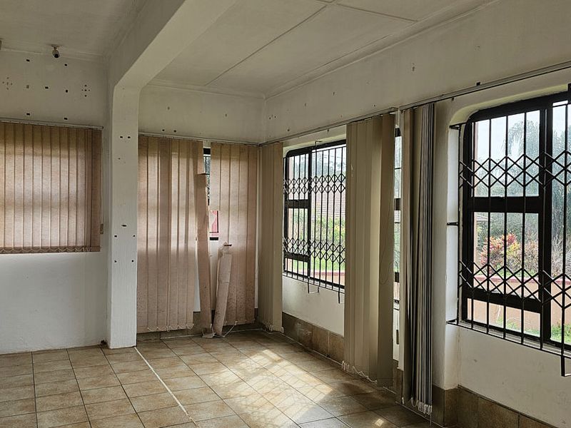An empty living area featuring tiled flooring, large windows with security gates, and vertical blinds, offering a bright space ready for renovation.