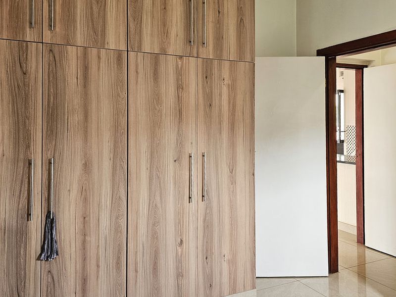 A bedroom featuring modern wood-grain built-in cupboards and neutral tiled flooring, leading into an adjacent hallway.