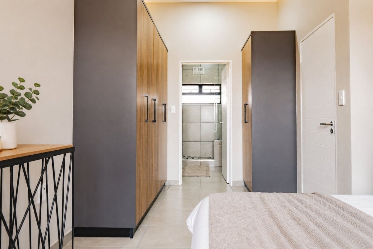 A modern bedroom featuring built-in cupboards with wood-grain and dark grey finishes, leading directly into an en-suite bathroom with tiled flooring.