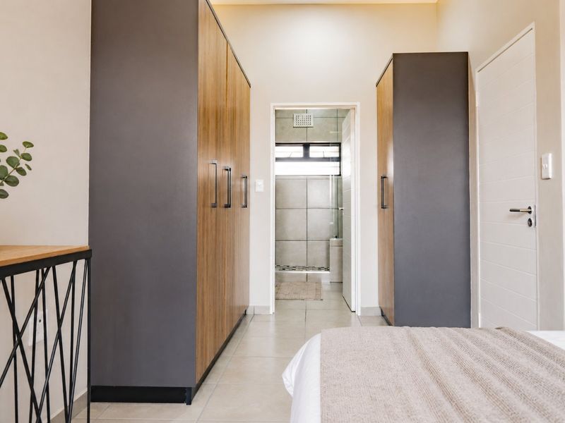 A modern bedroom featuring built-in cupboards with wood-grain and dark grey finishes, leading directly into an en-suite bathroom with tiled flooring.
