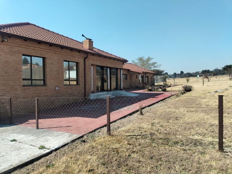 Rear view of a brick farmhouse featuring a paved patio area, sliding glass doors, and a tiled roof, set against a large open field.