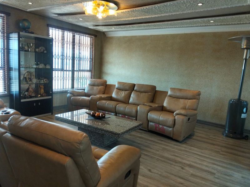 A spacious living room featuring laminate flooring, textured ceiling details with recessed lighting, and large windows fitted with security gates and blinds.