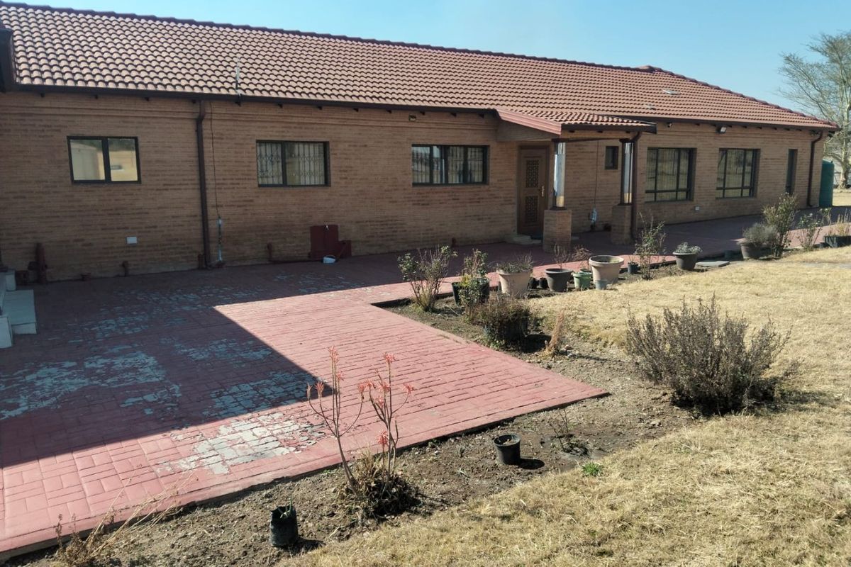 Rear view of a brick farmhouse featuring a large paved patio area and a tiled roof, set against a dry garden landscape.
