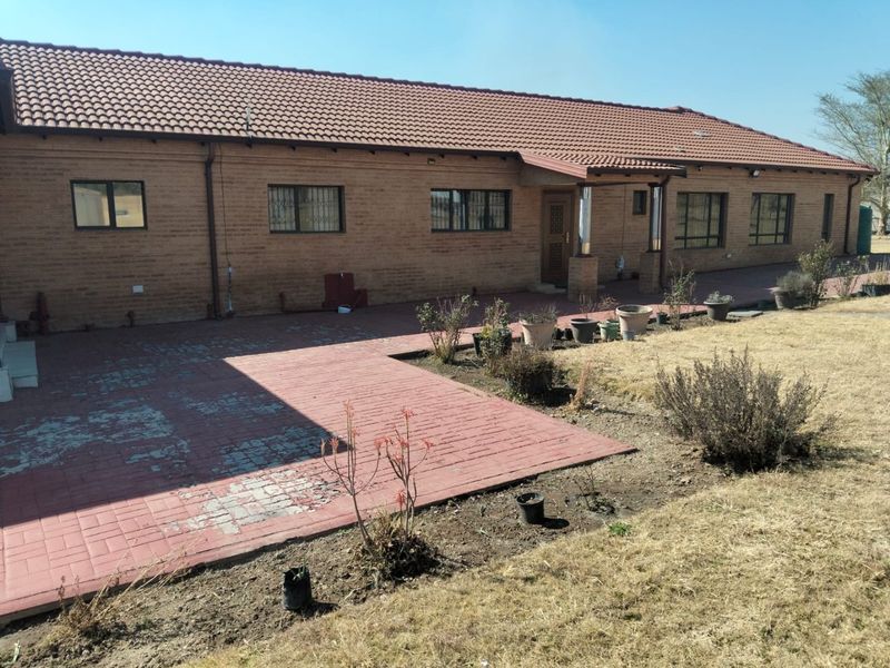 Rear view of a brick farmhouse featuring a large paved patio area and a tiled roof, set against a dry garden landscape.