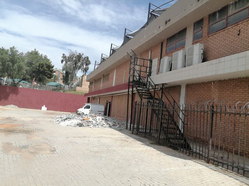 Rear exterior view of a commercial building featuring a brick facade, external metal staircase, security fencing, and a paved yard area with debris.