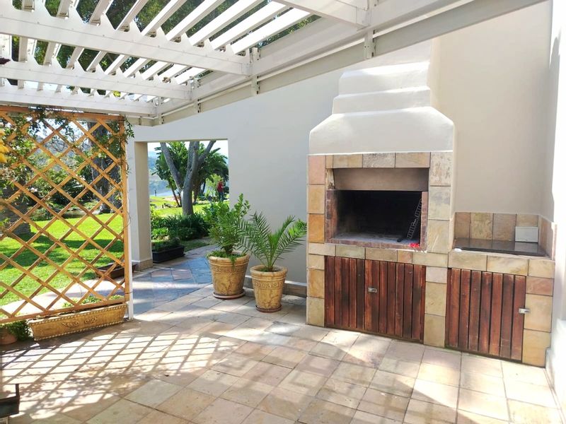 Covered outdoor patio featuring a built-in braai, tiled flooring, and a wooden pergola, overlooking a landscaped garden area.
