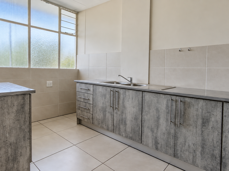 A clean, modern kitchen featuring grey wood-look cabinetry, laminate countertops, a stainless steel sink, and neutral tiled splashbacks with ample natural light.