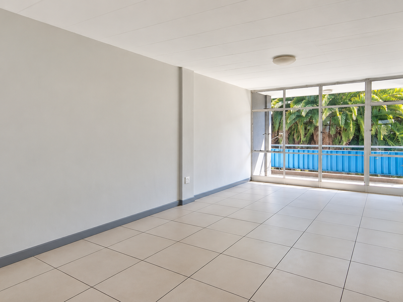 A bright, empty living area featuring neutral tiled flooring, clean white walls, and large windows providing natural light and a view of greenery.