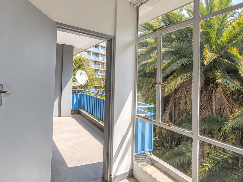 A bright, empty balcony area featuring large metal-framed windows and access to an outdoor walkway with a blue metal railing.