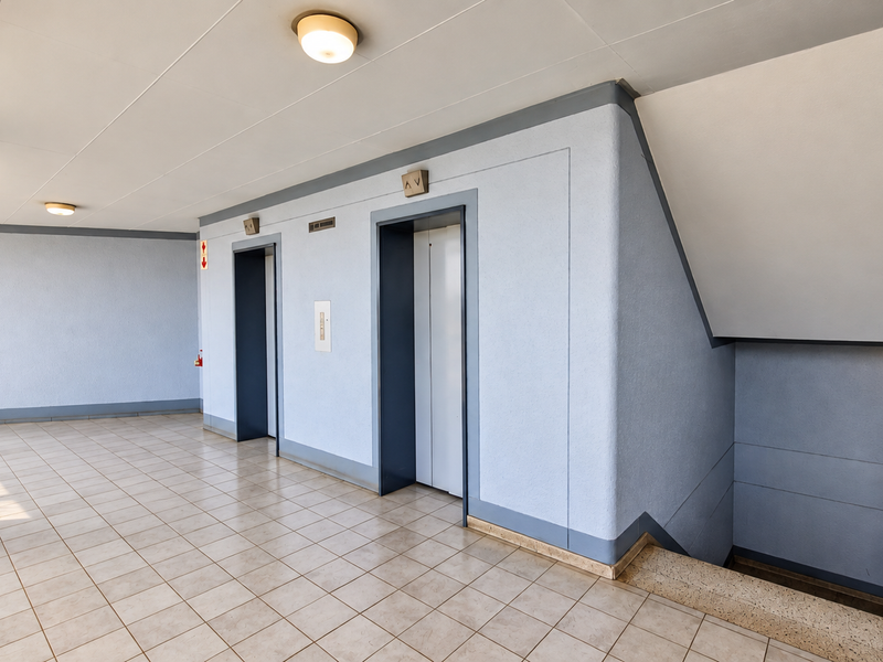 A clean and well-maintained apartment building hallway featuring elevator access, a staircase with metal railings, and neutral tiled flooring.