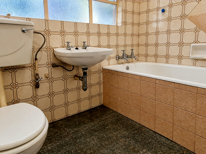 A dated bathroom featuring original ceramic wall tiles, a standard bathtub, pedestal basin, and flush toilet with exposed plumbing fixtures.