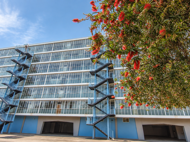 Exterior view of a multi-story apartment building featuring extensive glass window facades, external steel fire escape staircases, and ground-level covered parking.