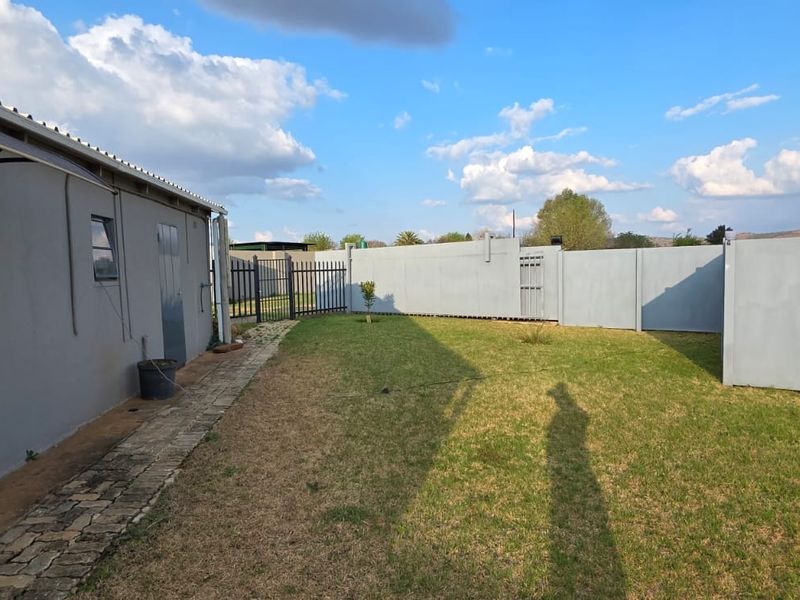 A rear exterior view of the property featuring a paved walkway, a lawn area, and secure perimeter walling with metal fencing.