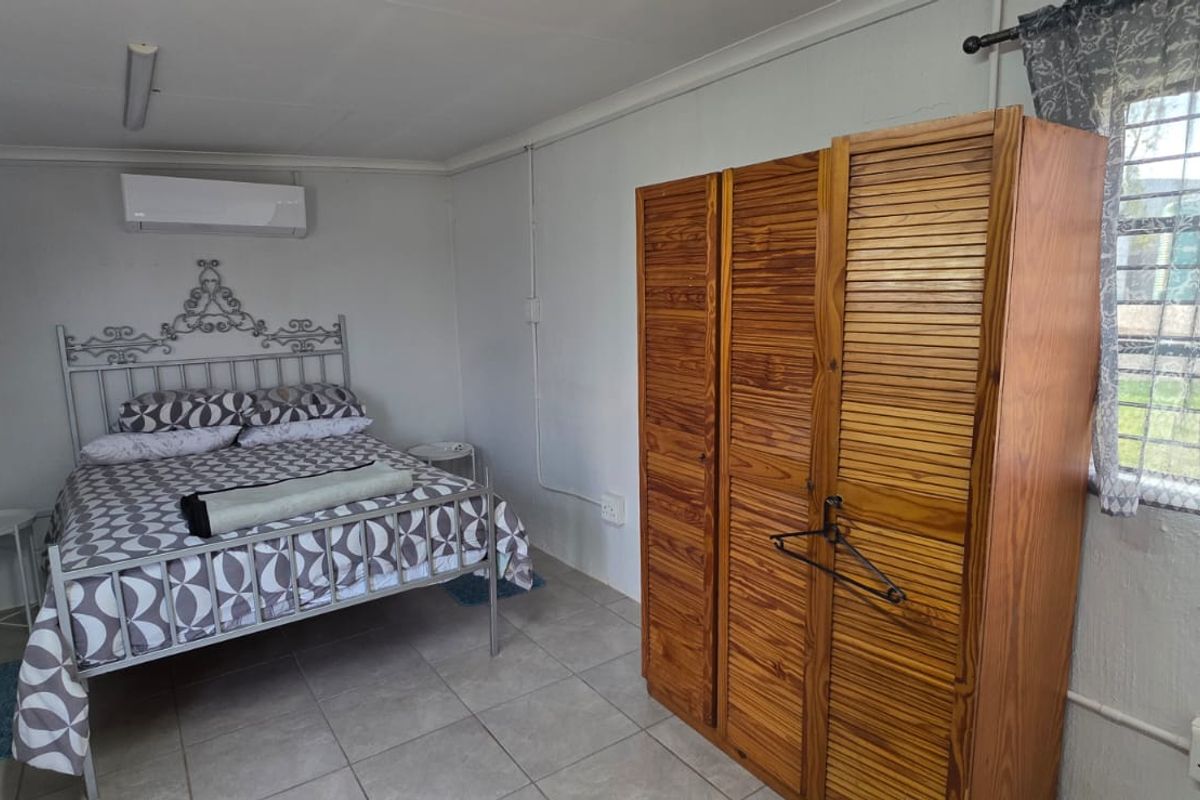 A functional bedroom featuring tiled flooring, a wall-mounted air conditioning unit, and windows fitted with security bars.