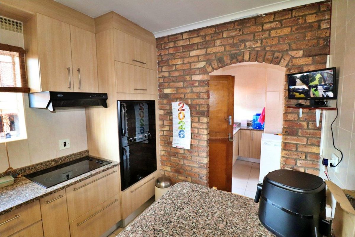 A functional kitchen featuring light wood cabinetry, granite countertops, an electric hob, and a distinctive exposed brick archway leading to an adjacent utility area.