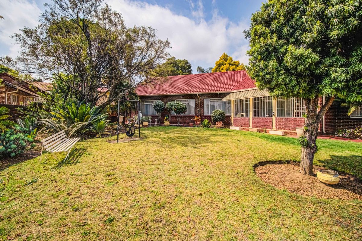 A spacious rear garden featuring a well-maintained lawn, mature trees, and a brick-built house with a tiled roof and covered patio area.