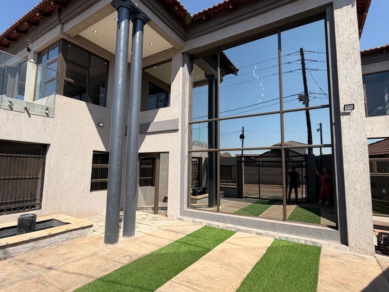 Modern double-storey rear exterior featuring a striking glass curtain wall, structural columns, paved patio with artificial lawn, and a built-in water feature.
