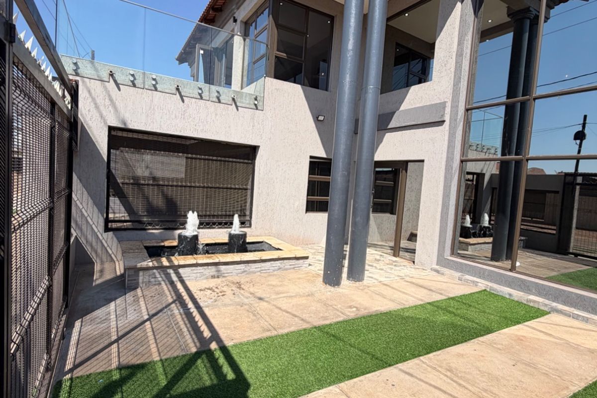 A modern rear exterior featuring a tiled patio, artificial lawn, a built-in water feature, and a striking double-story facade with glass balustrades.