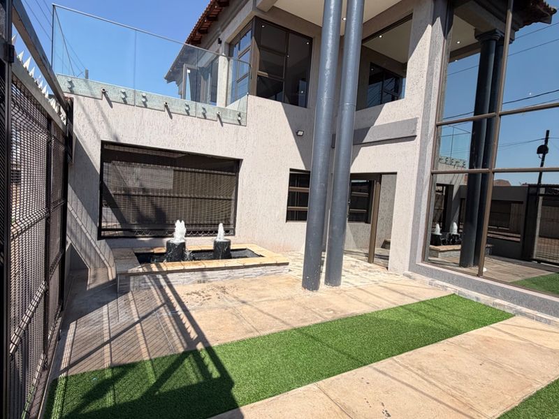 A modern rear exterior featuring a tiled patio, artificial lawn, a built-in water feature, and a striking double-story facade with glass balustrades.
