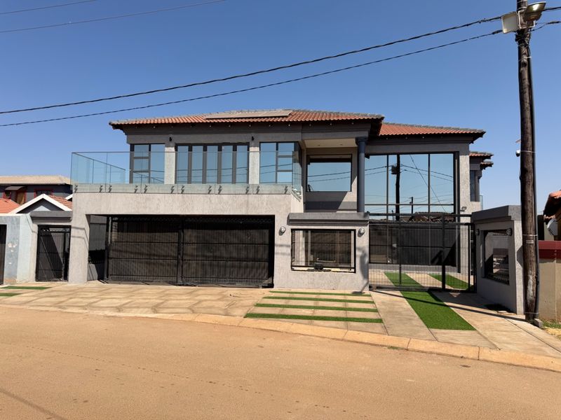 Modern double-story residential facade featuring a terracotta tile roof, glass balustrade balcony, large windows, and a paved driveway with integrated artificial grass.
