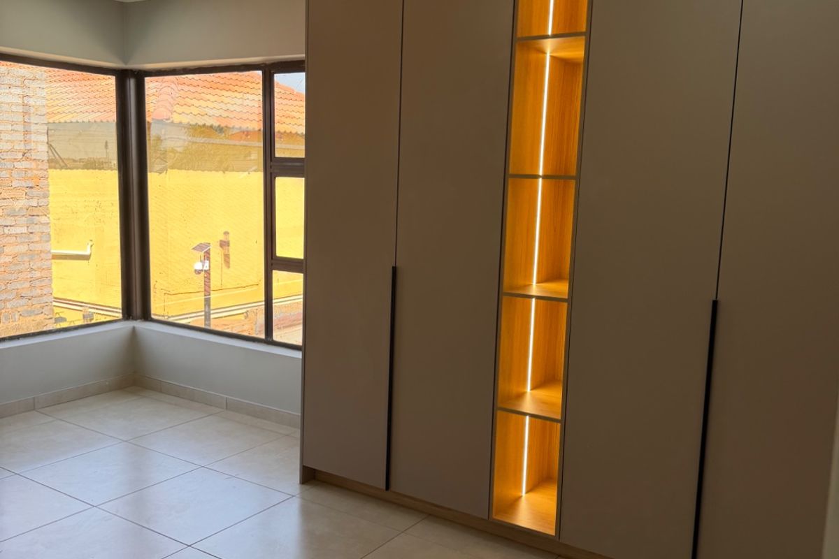 A bright, modern bedroom featuring large corner windows, neutral tiled flooring, and a contemporary built-in wardrobe with integrated LED display shelving.