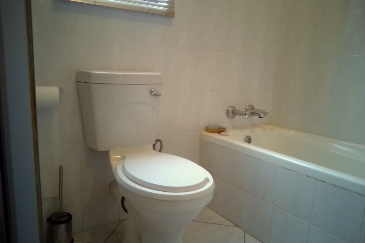 A functional bathroom featuring a standard toilet, a built-in bathtub with wall-mounted mixer taps, and neutral wall and floor tiling.
