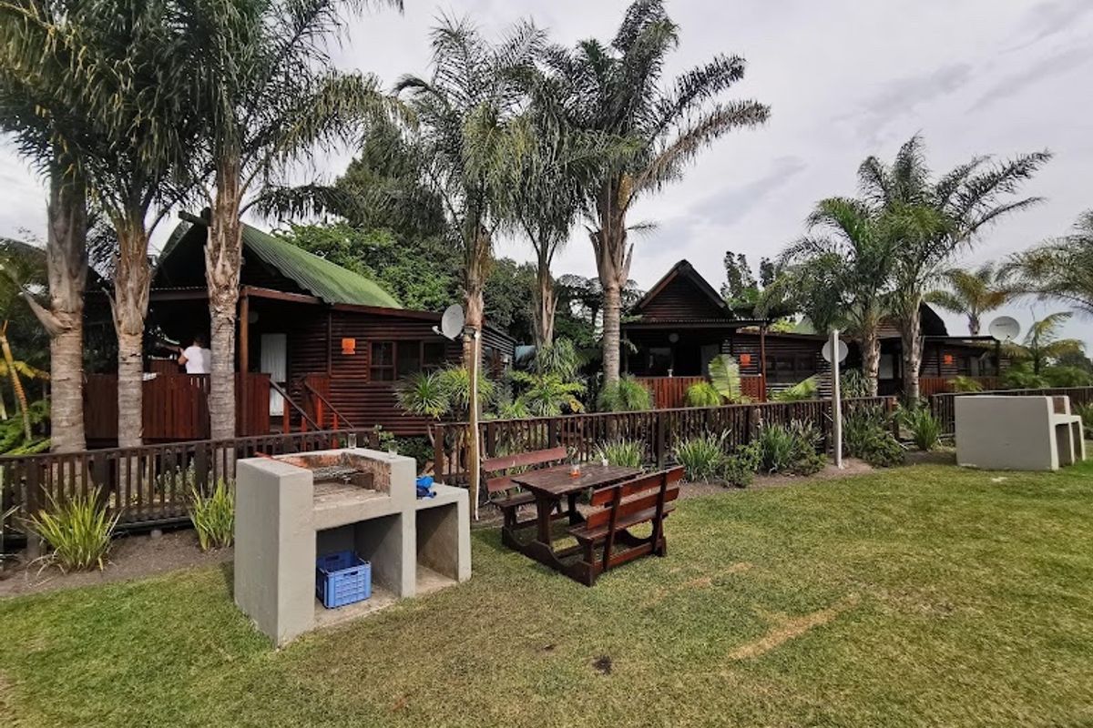 A row of timber cabins featuring elevated wooden decks, set within a landscaped garden area with built-in concrete braai facilities.