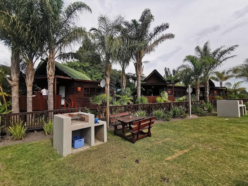 A row of timber cabins featuring elevated wooden decks, set within a landscaped garden area with built-in concrete braai facilities.