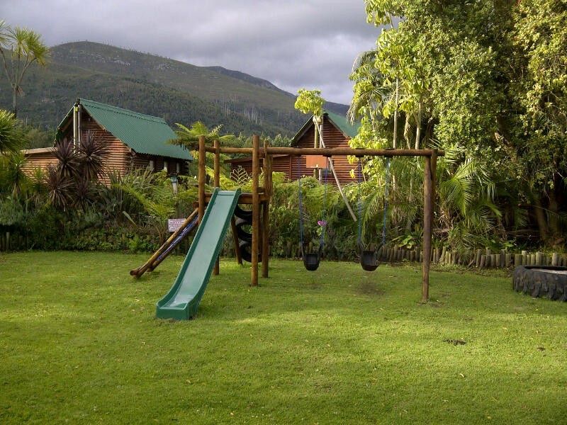 A well-maintained garden area featuring a wooden playground structure with a slide and swings, set against a backdrop of timber cabins and mountain views.