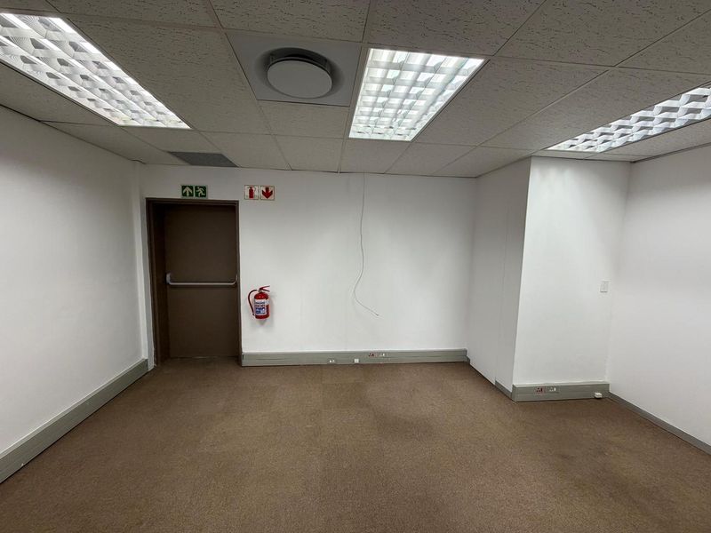 An empty office space featuring a suspended ceiling with recessed lighting, carpeted flooring, and an emergency exit door.