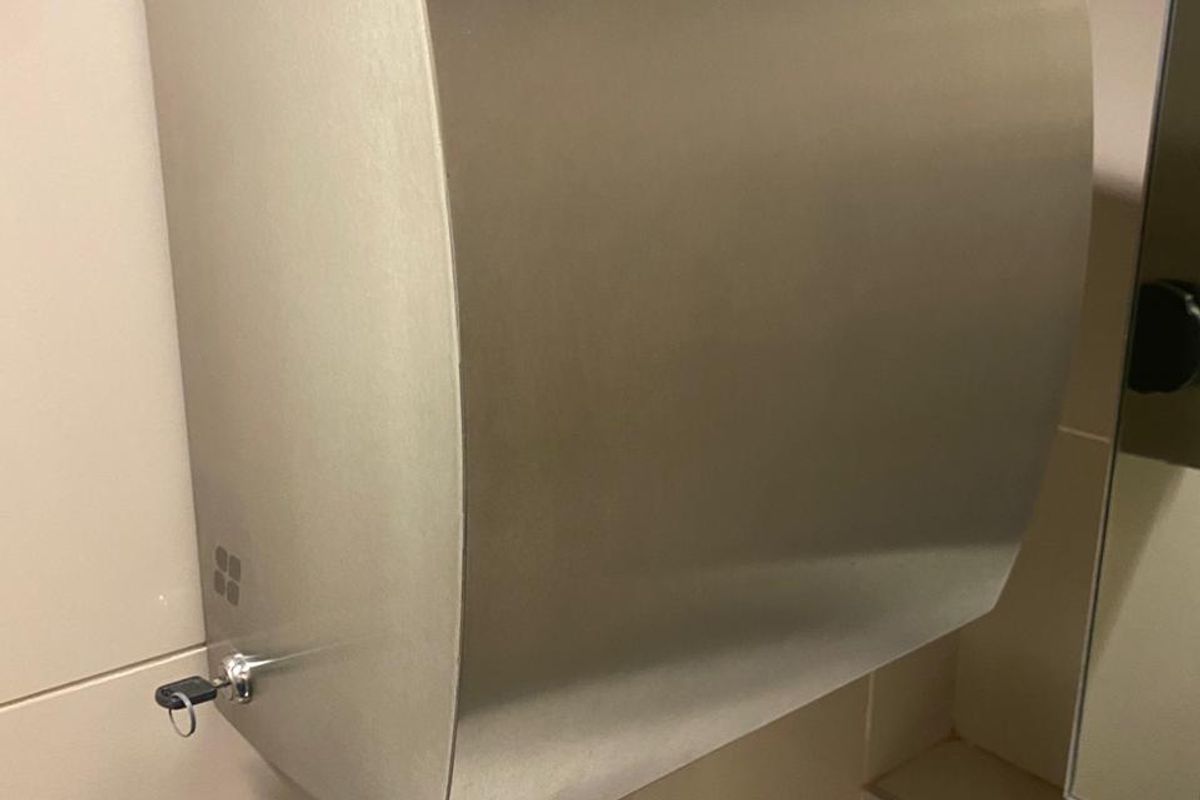 A close-up view of a wall-mounted stainless steel automatic paper towel dispenser installed in a tiled bathroom facility.