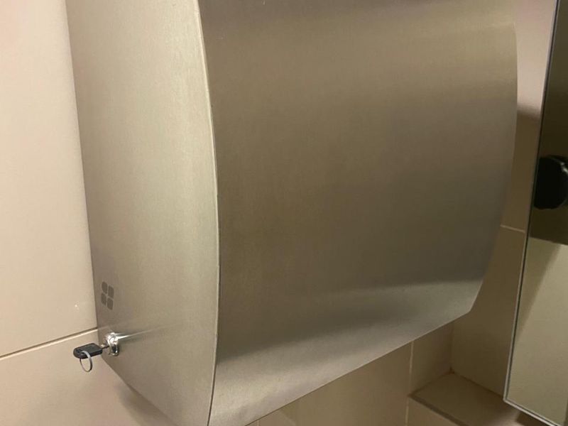 A close-up view of a wall-mounted stainless steel automatic paper towel dispenser installed in a tiled bathroom facility.