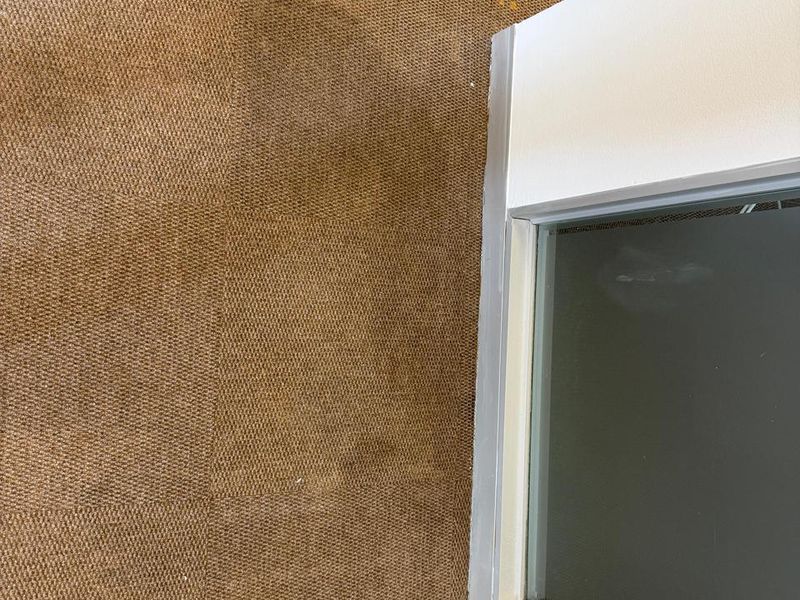 A close-up view of office flooring featuring brown carpet tiles adjacent to a glass partition with an aluminum frame.