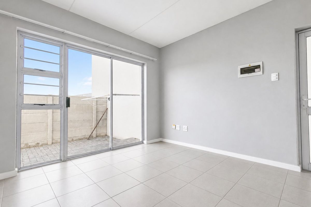 A bright, empty living area featuring neutral tiled flooring, a sliding glass door leading to an outdoor space, and a wall-mounted distribution board.
