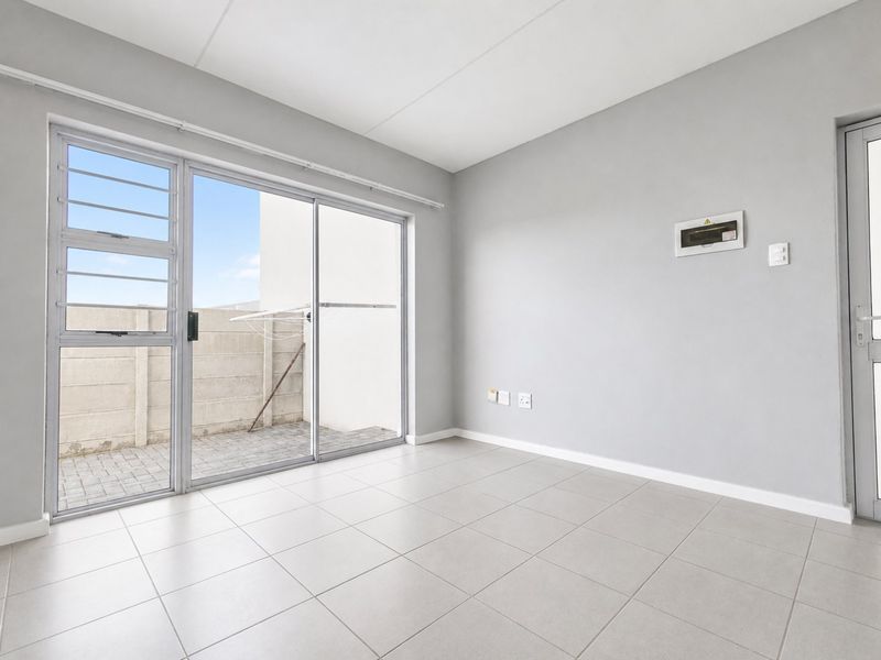 A bright, empty living area featuring neutral tiled flooring, a sliding glass door leading to an outdoor space, and a wall-mounted distribution board.