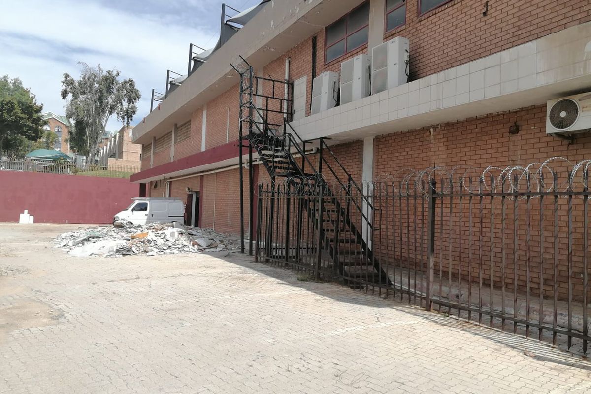 Rear exterior view of a commercial building featuring a brick facade, external metal staircase, security fencing, and paved parking area with visible debris.