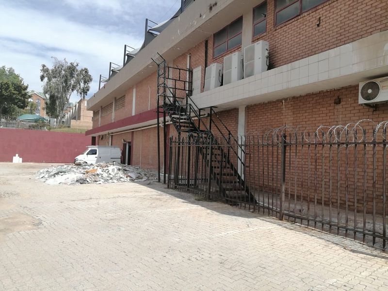 Rear exterior view of a commercial building featuring a brick facade, external metal staircase, security fencing, and paved parking area with visible debris.