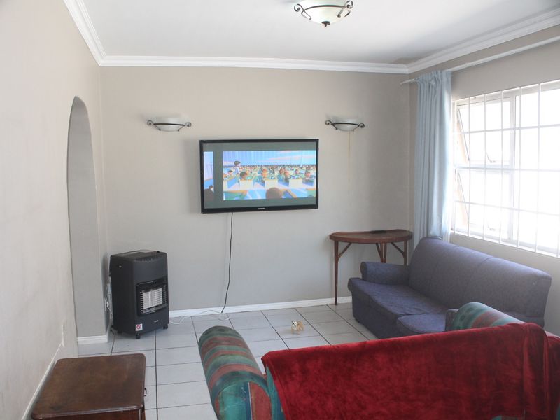 A living room featuring tiled flooring, wall-mounted lighting, and a large window with security bars, providing a functional and neutral living space.