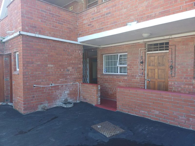 Exterior view of an apartment entrance featuring exposed brickwork, a timber door with security gate, and a paved driveway area.