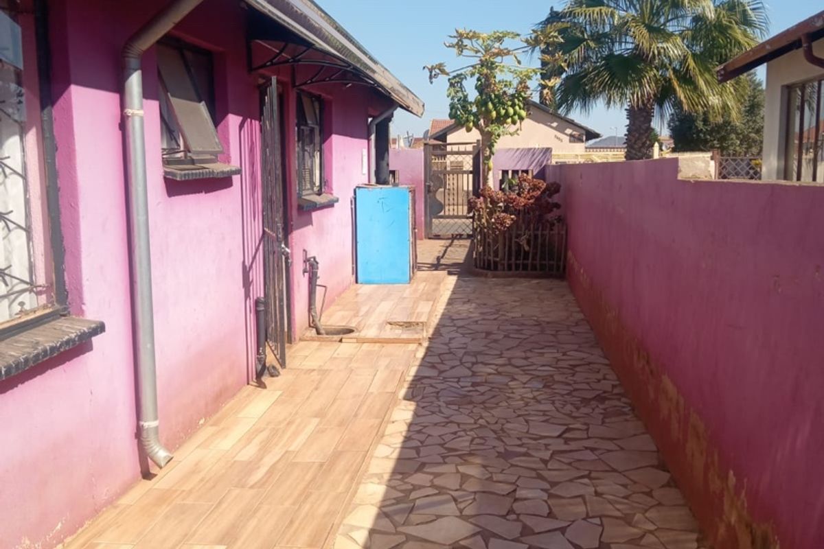A narrow rear exterior walkway featuring a combination of wood-look tiles and stone paving, bordered by a boundary wall with visible maintenance requirements.