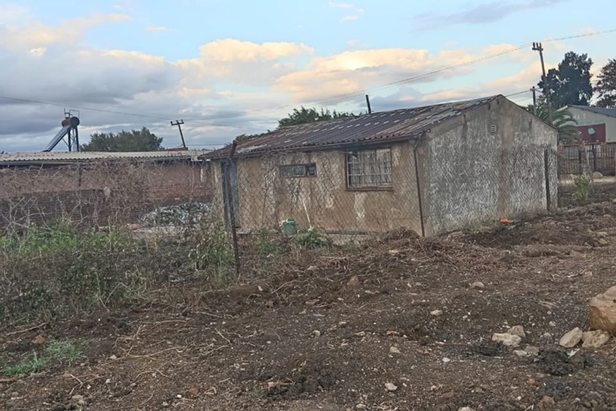 A detached structure with a corrugated metal roof and security bars on windows, situated on an undeveloped plot of land.