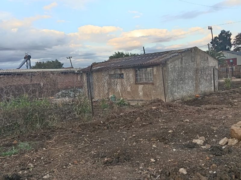 A detached structure with a corrugated metal roof and security bars on windows, situated on an undeveloped plot of land.
