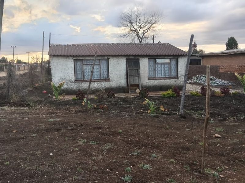 A modest single-story detached house featuring a corrugated metal roof and security bars on the windows, set on an unpaved plot.