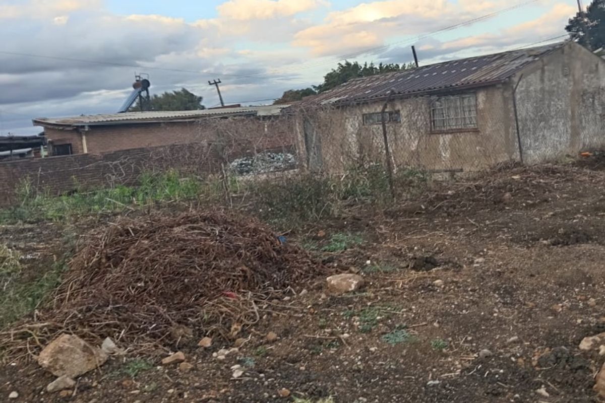 Rear view of the property showing a detached structure with masonry walls, corrugated roofing, and security bars, set on an undeveloped plot.