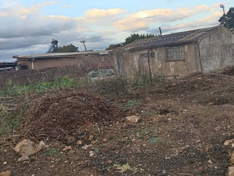Rear view of the property showing a detached structure with masonry walls, corrugated roofing, and security bars, set on an undeveloped plot.