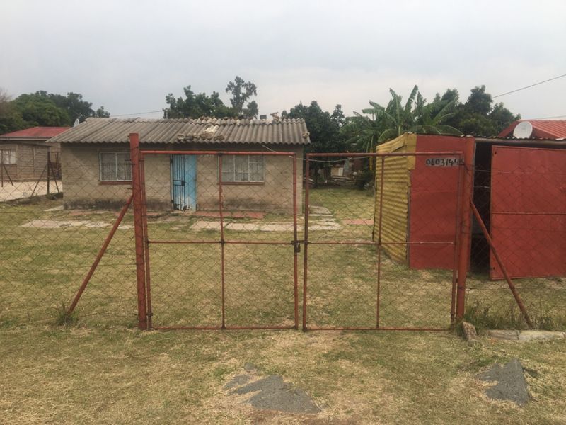 Front view of a modest single-story dwelling featuring a corrugated roof, concrete block construction, and a fenced yard with a metal gate.