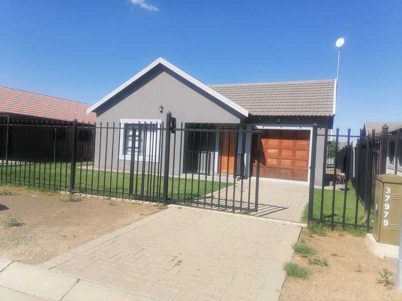 A well-maintained single-story house featuring a grey facade, tiled roof, wooden garage door, and secure metal perimeter fencing with a paved driveway.