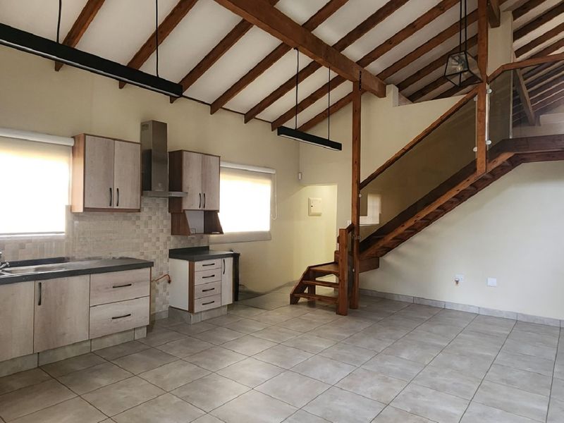 Spacious open-plan living area featuring a modern kitchen, tiled floors, exposed timber rafters, and a striking timber staircase with glass balustrade.