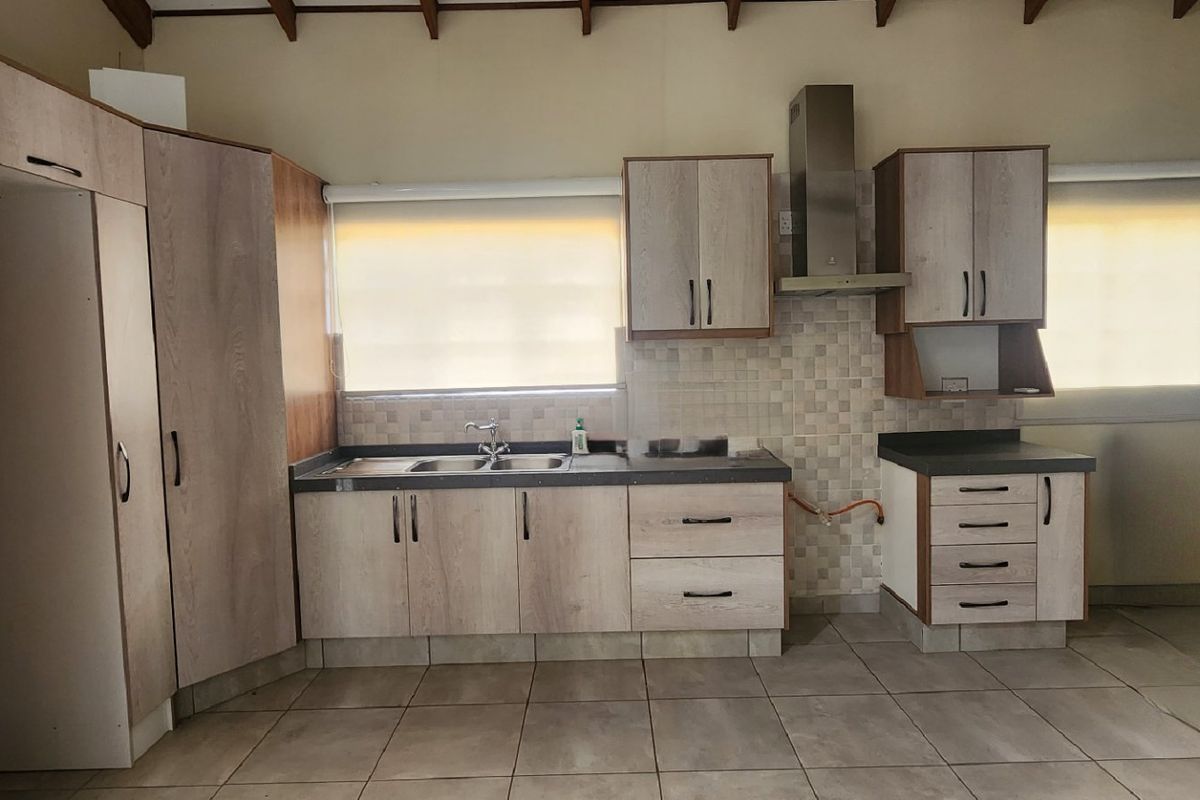 A functional kitchen featuring light wood-look cabinetry, a stainless steel extractor fan, tiled backsplash, and exposed timber ceiling beams in an open-plan layout.